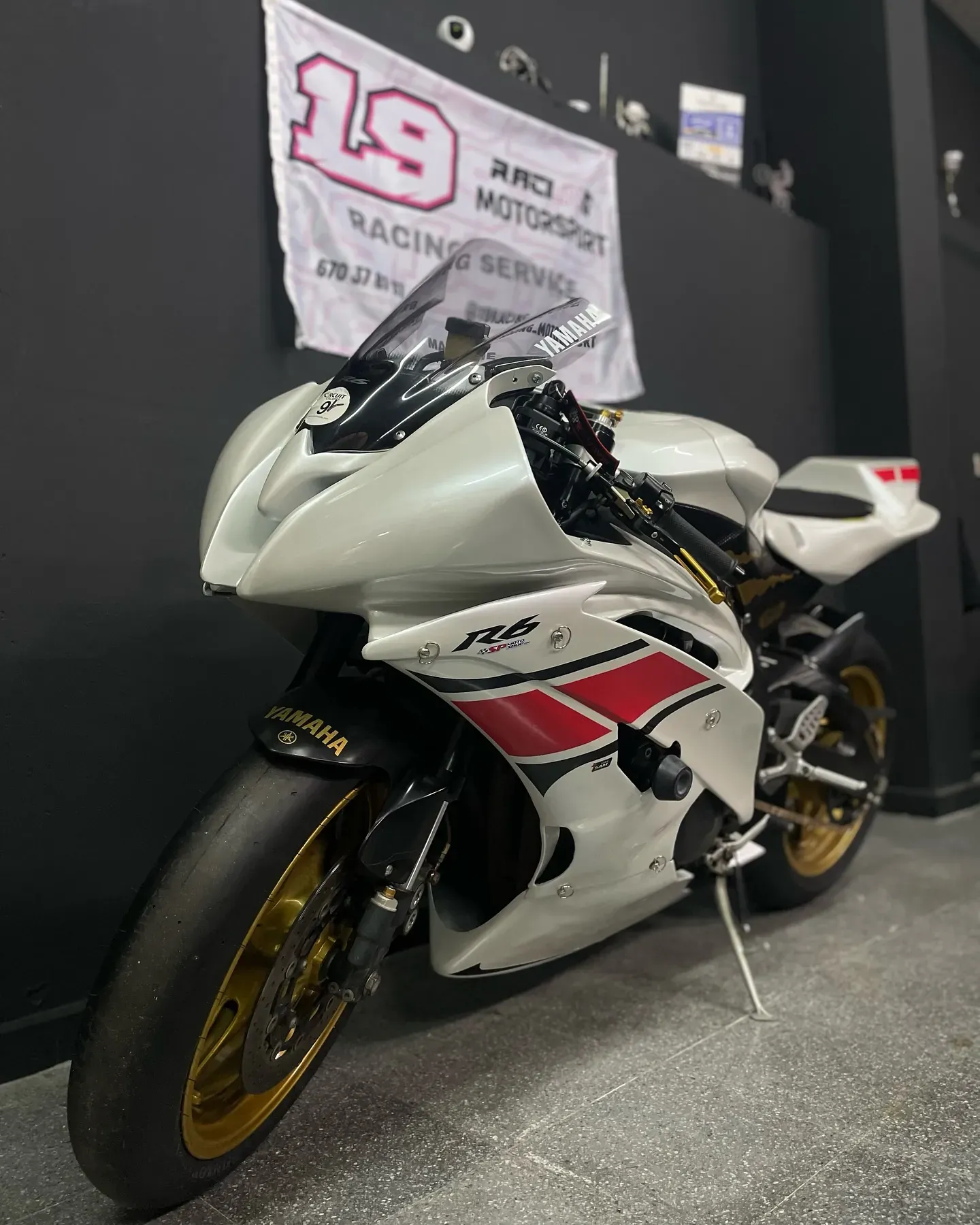 Yamaha R6 blanca y roja con llantas doradas en el taller, bajo la bandera de 19 Racing Motorsport